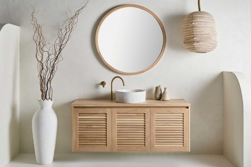 An alura round mirror in a bathroom with a keys range freestanding vanity with a stone sink. The neutral bathroom is styled with a low-hanging light and a white pot with twisted willow.