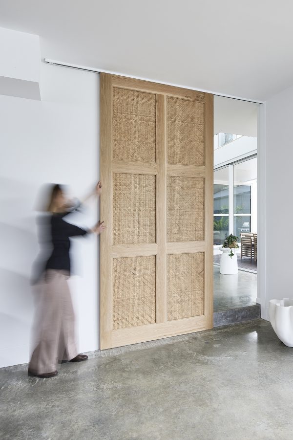 A blurred female image pushing closed the internal Pacific Door in a home. The door is custom made from Light American Oak with 6 open weave rattan panels