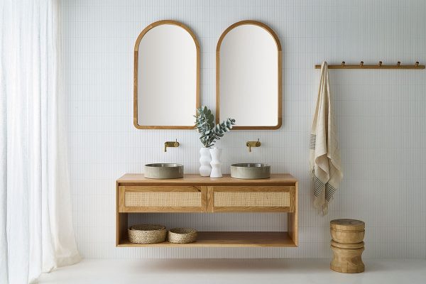 A Pacific Baxter Vanity wall hung with an open shelf and two drawers with rattan profiles. This wall hung vanity is in a coastal bathroom setting with two stone sinks, brass tapware and under two alura mirrors.