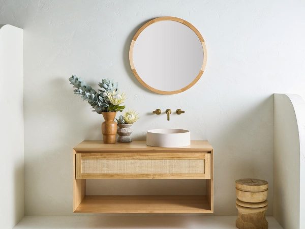 The Pacific Baxter Vanity, a custom made timber vanity with a bottom open shelf and rattan drawer profile with a single stone sink, brass tapware and an Alura Round mirror in a coastal bathroom setting